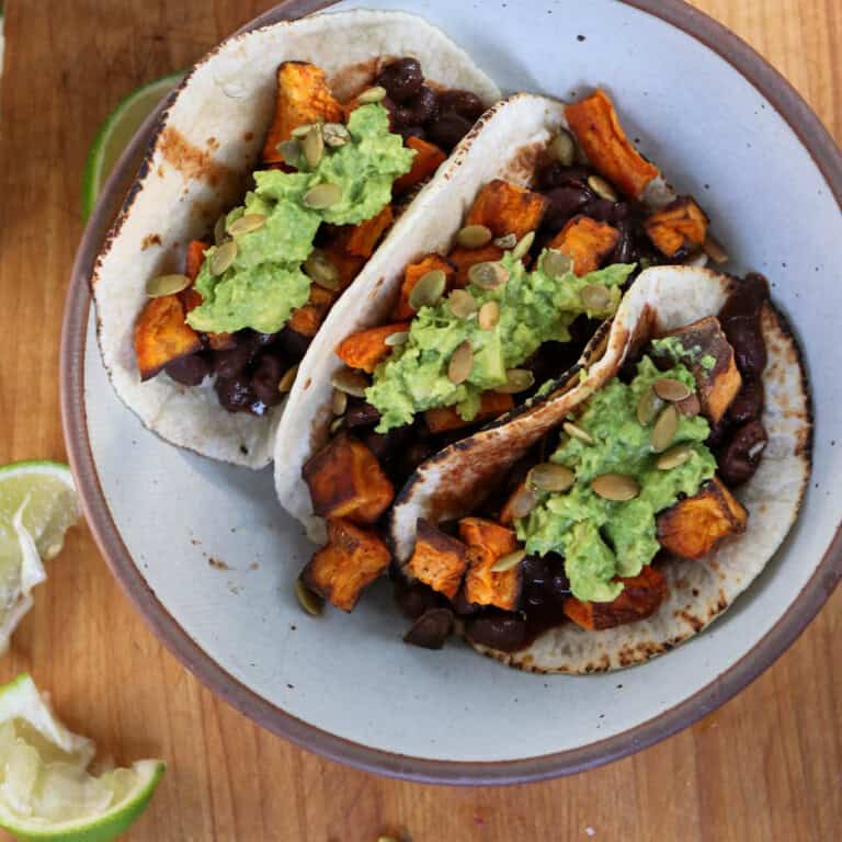 Sweet potato and black bean tacos, guacamole, and garnished with pumpkin seeds. The bowl is placed on a wooden surface, with lime wedges visible beside it.