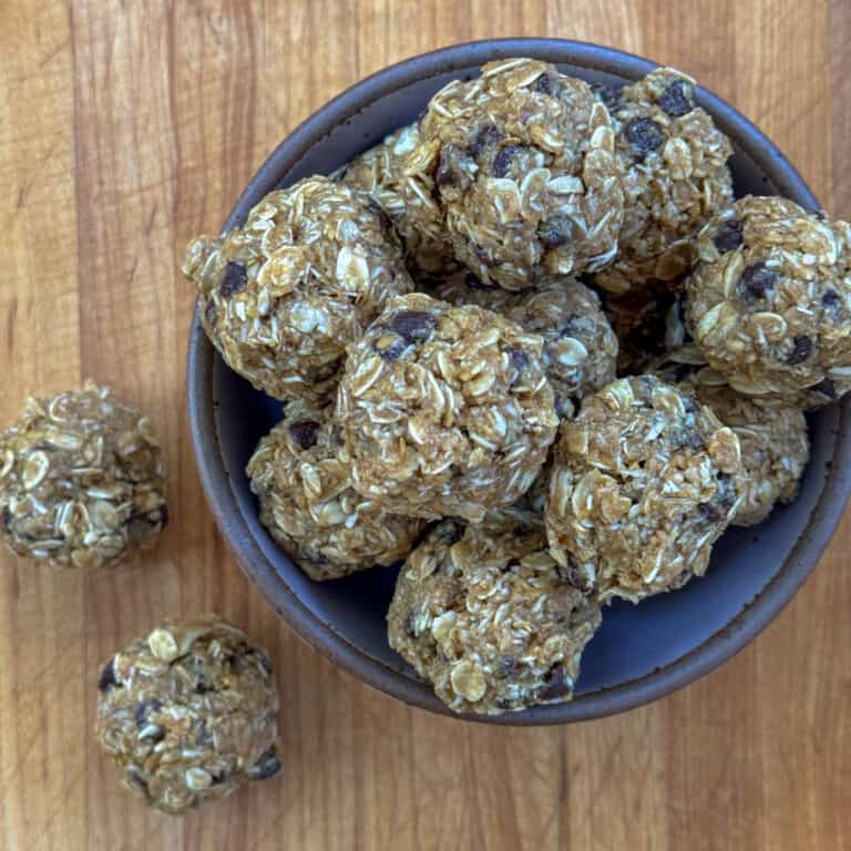 A top-down view of a bowl filled with homemade no-bake energy bites made with oats, chocolate chips, and other ingredients. Three additional energy bites are placed on the wooden surface beside the bowl. The energy bites have a slightly rough texture.