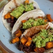 Three black bean and sweet potato tacos rest on a blue plate, brimming with guacamole and topped with crunchy pumpkin seeds. A lime wedge peeks out in the background.