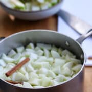 A pot filled with chopped green apples and a cinnamon stick, ready for cooking. A cutting board with a knife and another bowl of sliced apples are in the background on a wooden table.