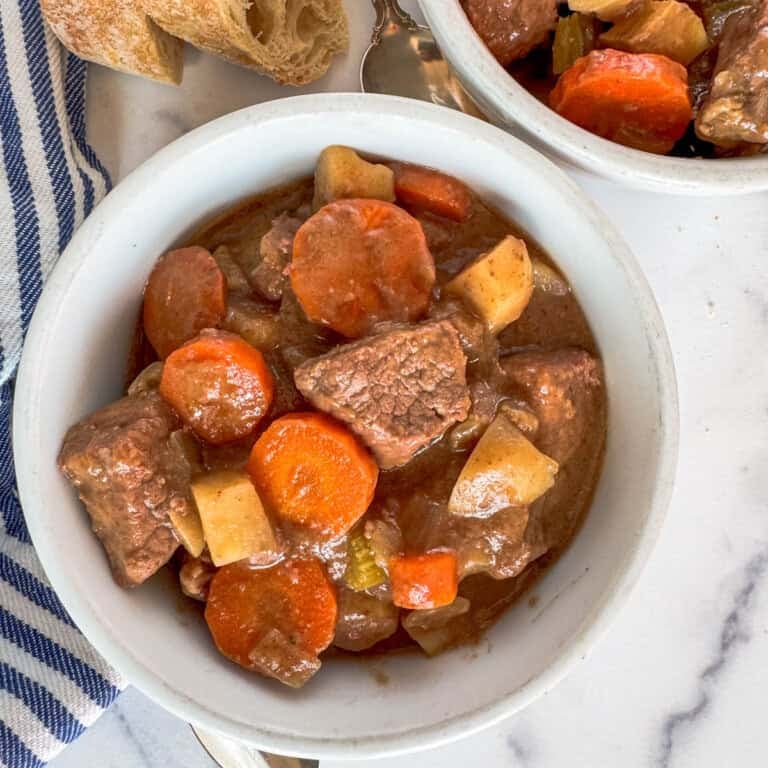 A bowl of slow cooker easy beef stew with chunks of beef, sliced carrots, and potatoes in a rich brown broth, served on a marble surface next to a piece of bread and a striped cloth.