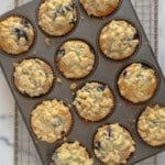 A dozen freshly baked blueberry muffins with crumb topping sit in a muffin tin on a cooling rack. The muffins are golden and slightly uneven, with visible blueberries and crumbly bits scattered across the tops.