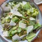 A fresh green salad in a bowl topped with shaved parmesan cheese, pistachios, and flaky sea salt, with two serving forks on the sides.