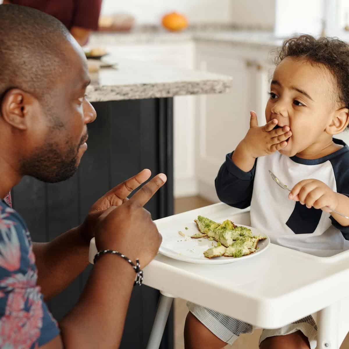An adult talks to a young child sitting in a high chair, who is eating avocado toast using their hand. In the bright, modern kitchen, they explore picky eating solutions together during mealtime.