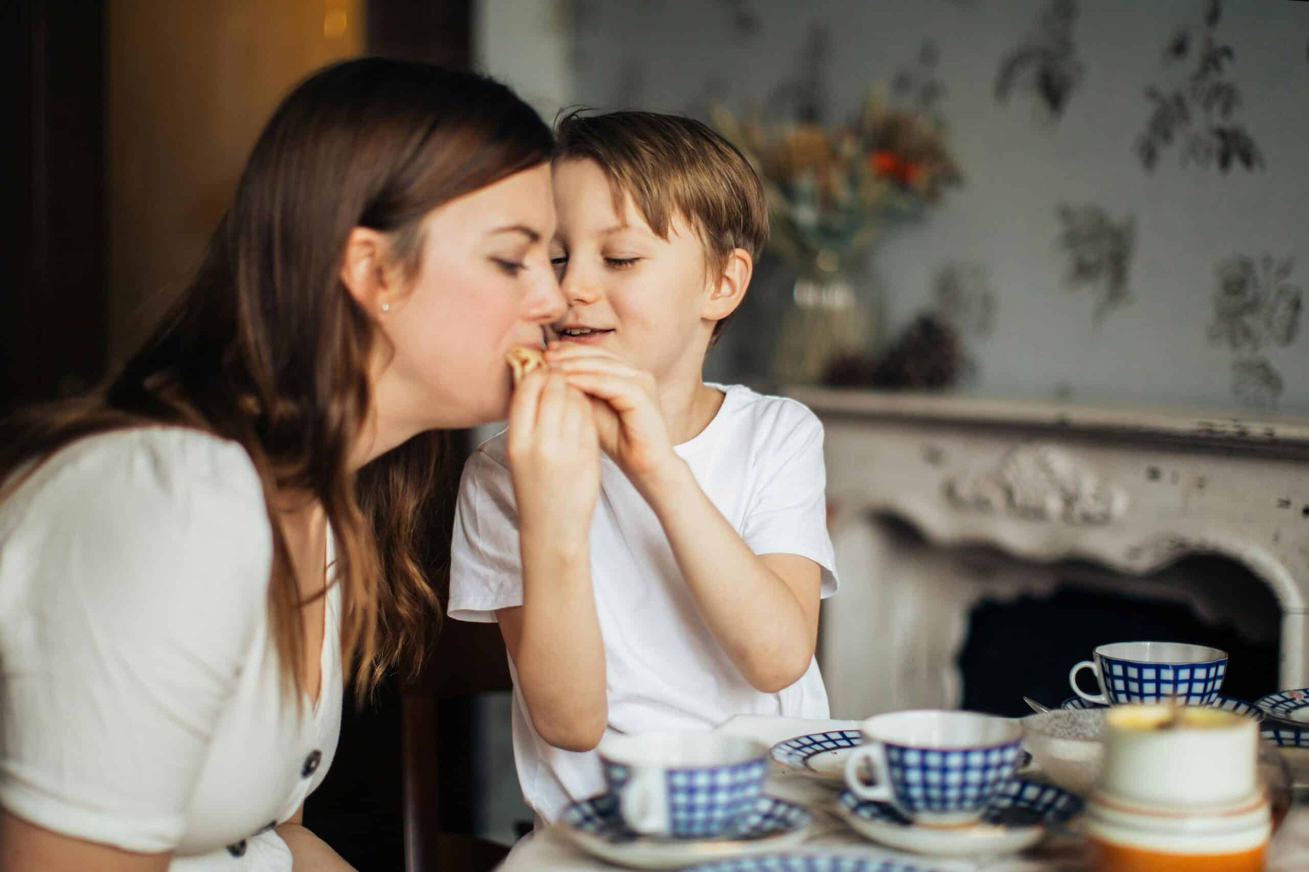 A young boy playfully sneaking food to a woman, likely his mother, as they sit together at a table set with tea and blue-striped cups in a cozy, warmly lit room—a charming moment of parenting and child behavior.