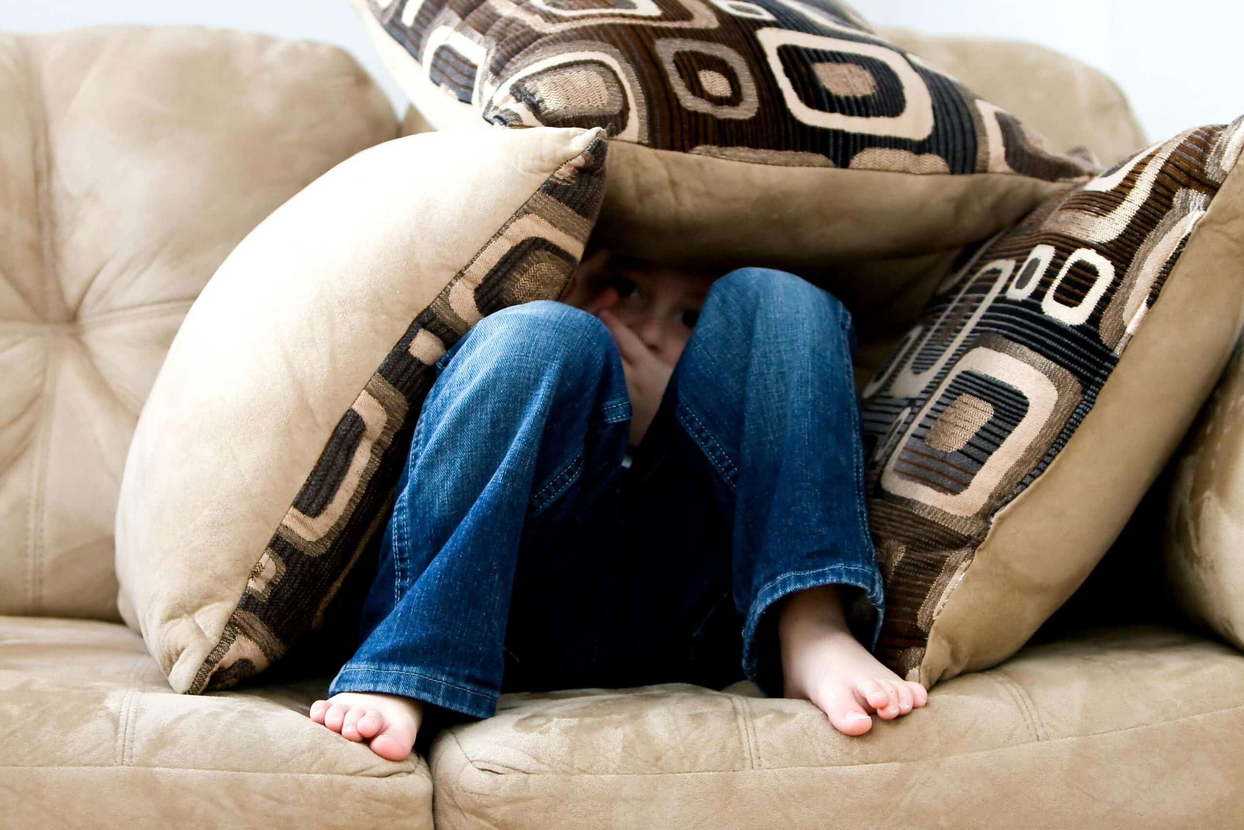 A child sits on a beige couch, mostly hidden under large patterned pillows, with only bare feet, legs in jeans, and part of their face visible—a playful moment that captures classic child behavior.