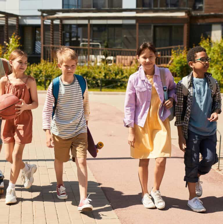 Four children walk outside together, two girls and two boys. One girl holds a basketball, another wears a yellow dress. One boy has a skateboard, and the other wears glasses—they're smiling, enjoying a sunny day, and are getting read for after school snacks as they laugh.