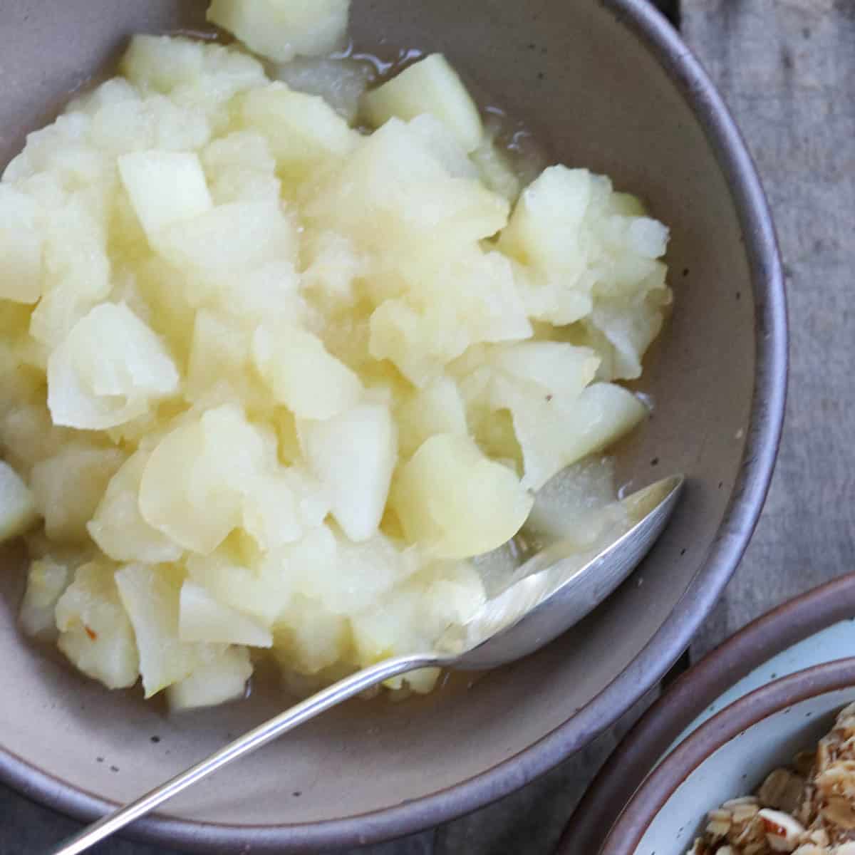 A bowl filled with chunky stovetop cooked apples, served with a spoon resting on the edge of the bowl. Another dish with granola is partially visible nearby.