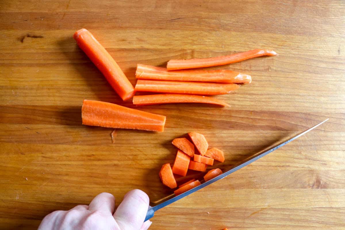 A hand is holding a knife, slicing a carrot for mixed green salad on a wooden cutting board.