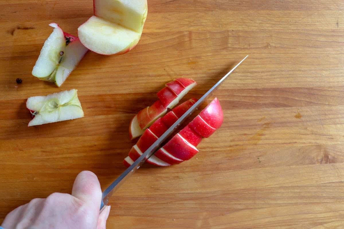 A hand uses a knife to slice a red apple on a wooden cutting board. The apple has already been cored and partially sliced into wedges and chopped for everyday salad.