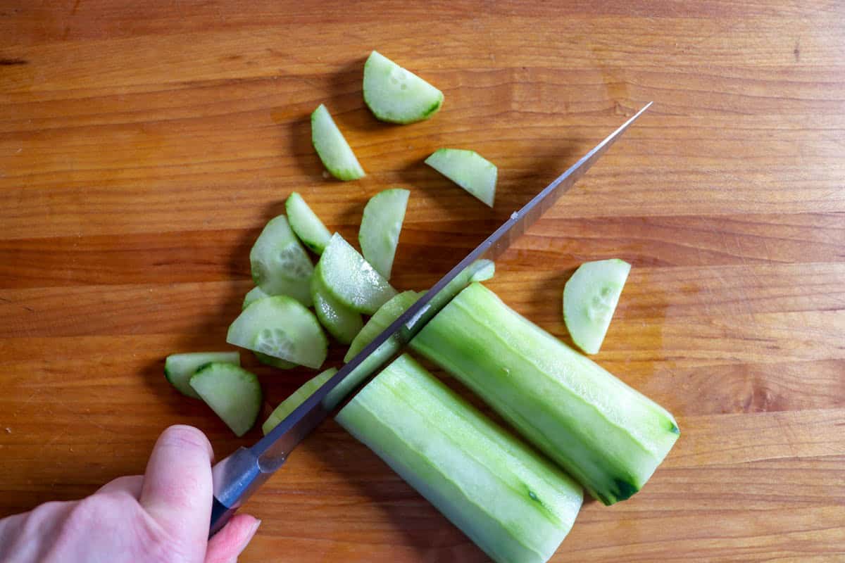 A hand is slicing a peeled cucumber on a wooden cutting board to make kids salad.