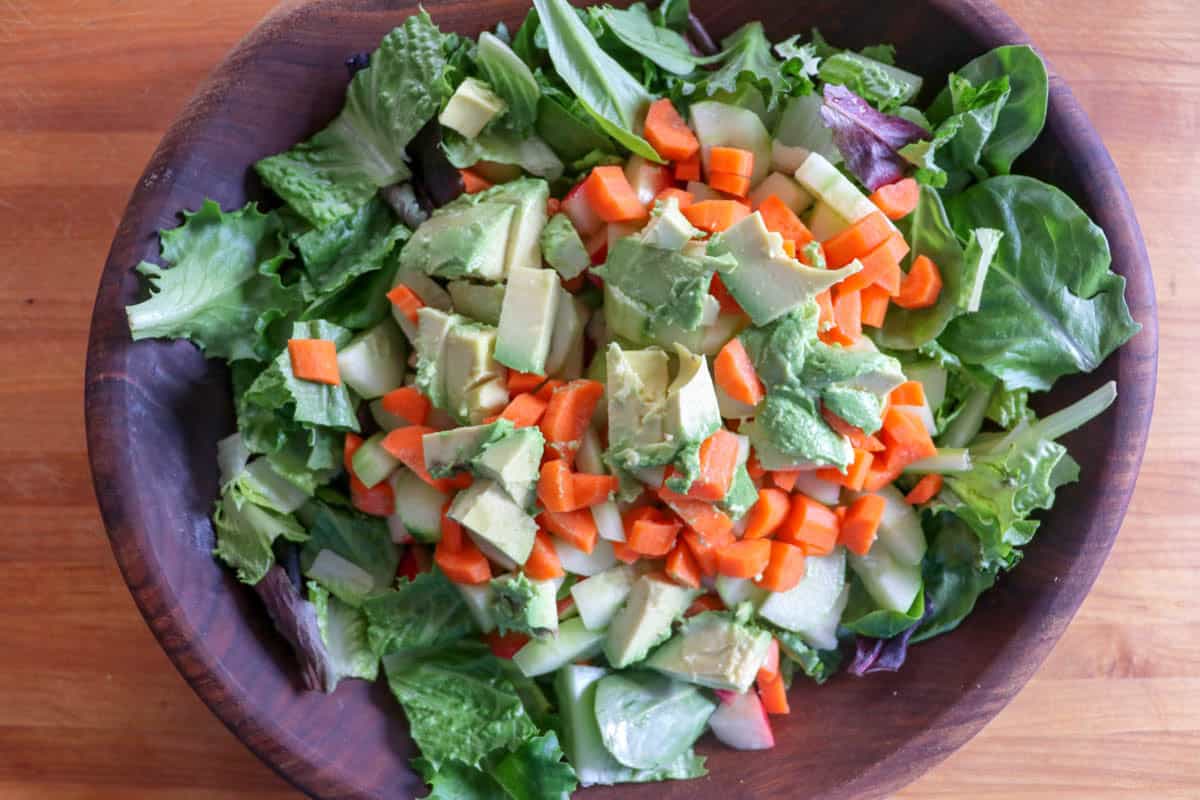 A wooden bowl filled with mixed greens, chopped carrots, diced avocado, and cucumber in a wooden bowl.