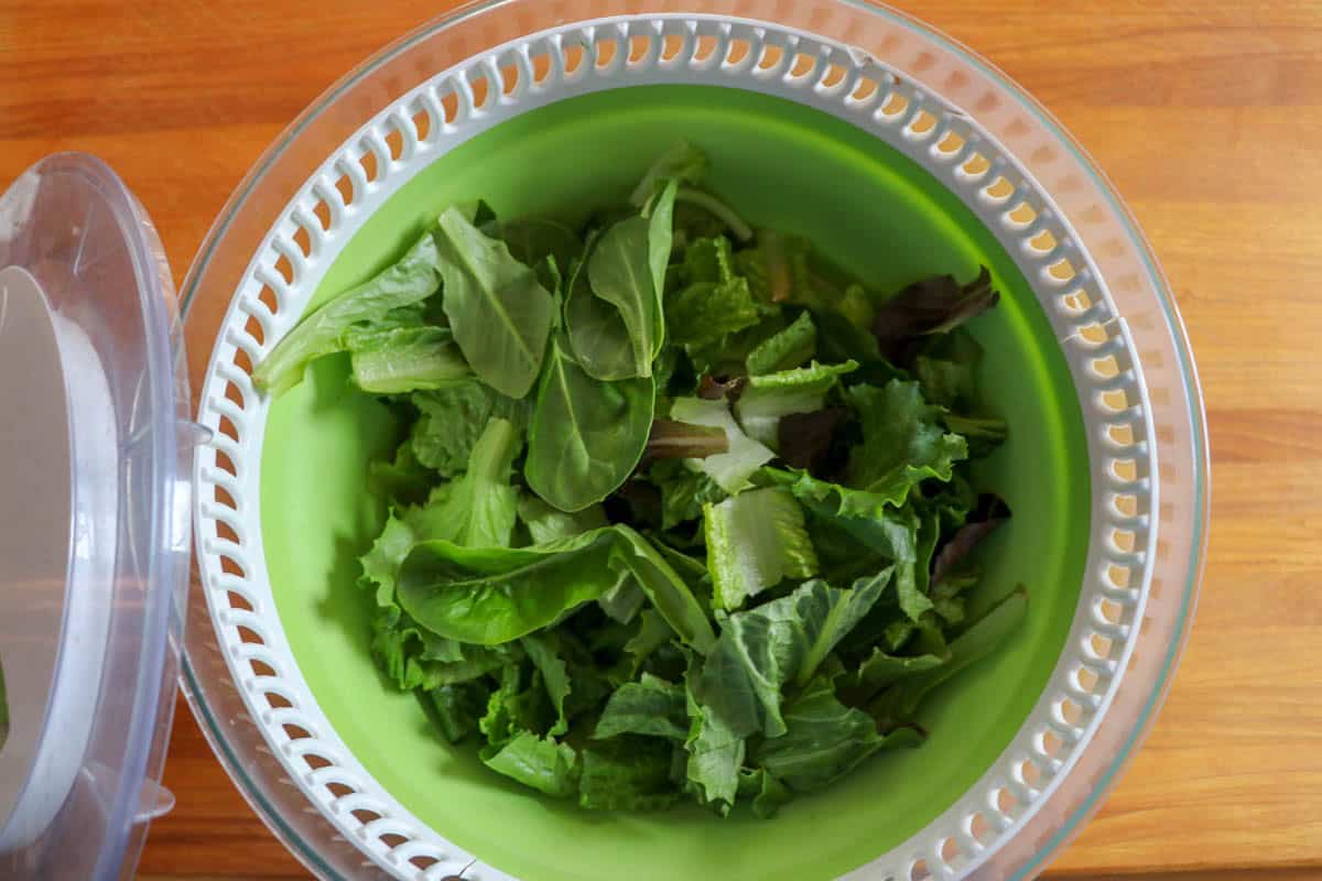 A top-down view of a fresh mixed green salad featuring lettuce leaves inside a green salad spinner, placed on a wooden surface.