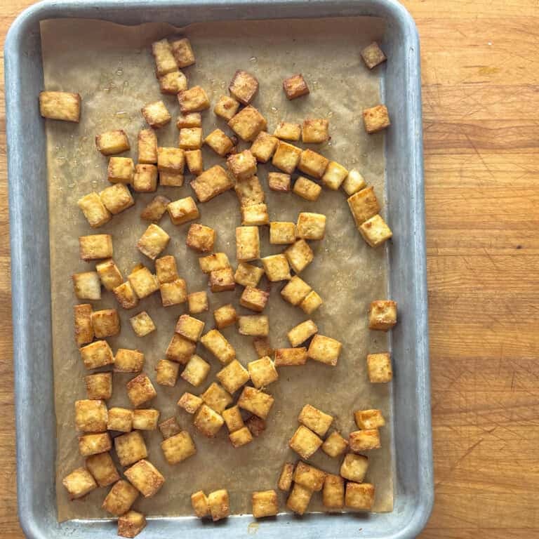 A baking sheet lined with parchment paper holds scattered, golden-brown simple baked tofu cubes on a wooden surface.