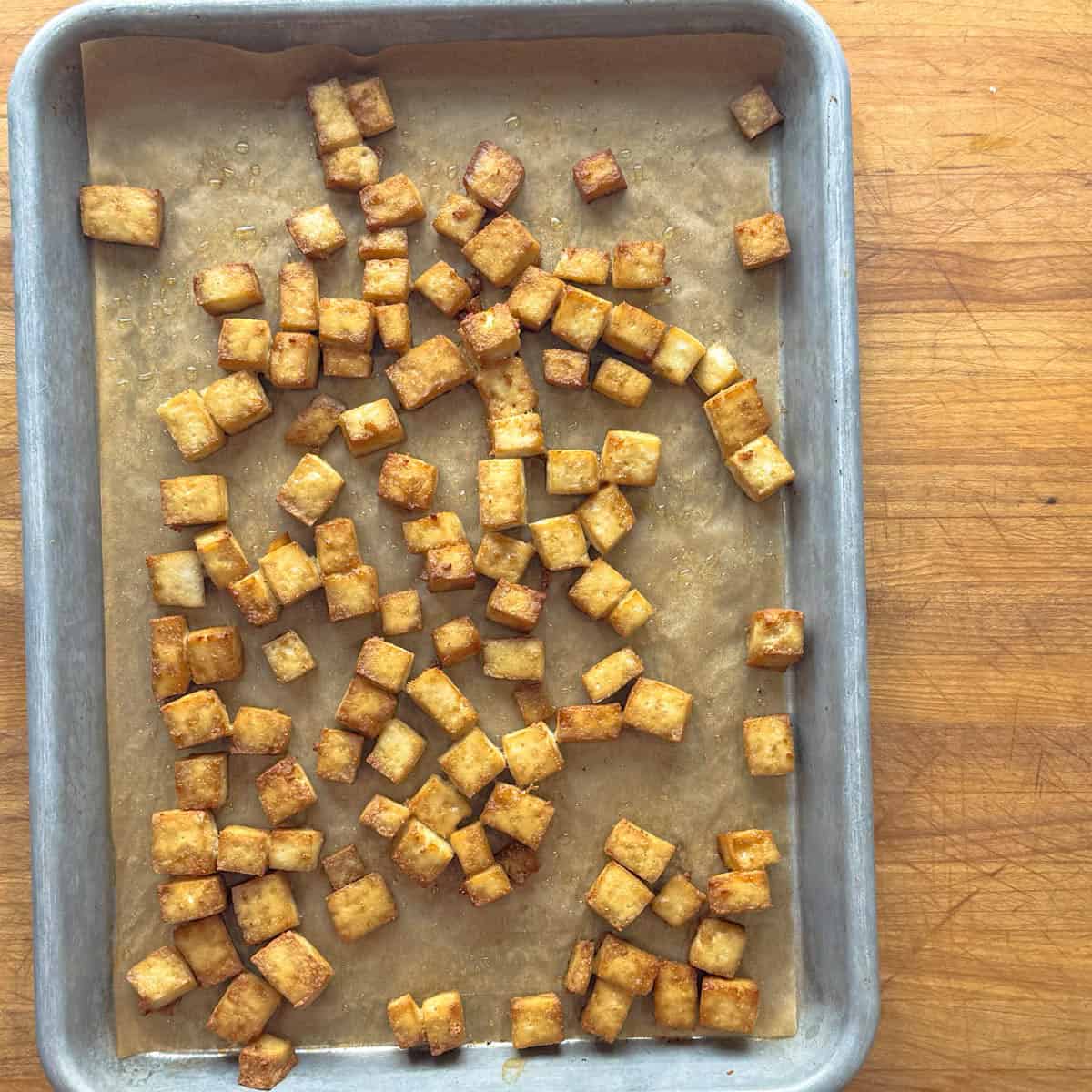 A baking sheet lined with parchment paper holds scattered, golden-brown simple baked tofu cubes on a wooden surface.