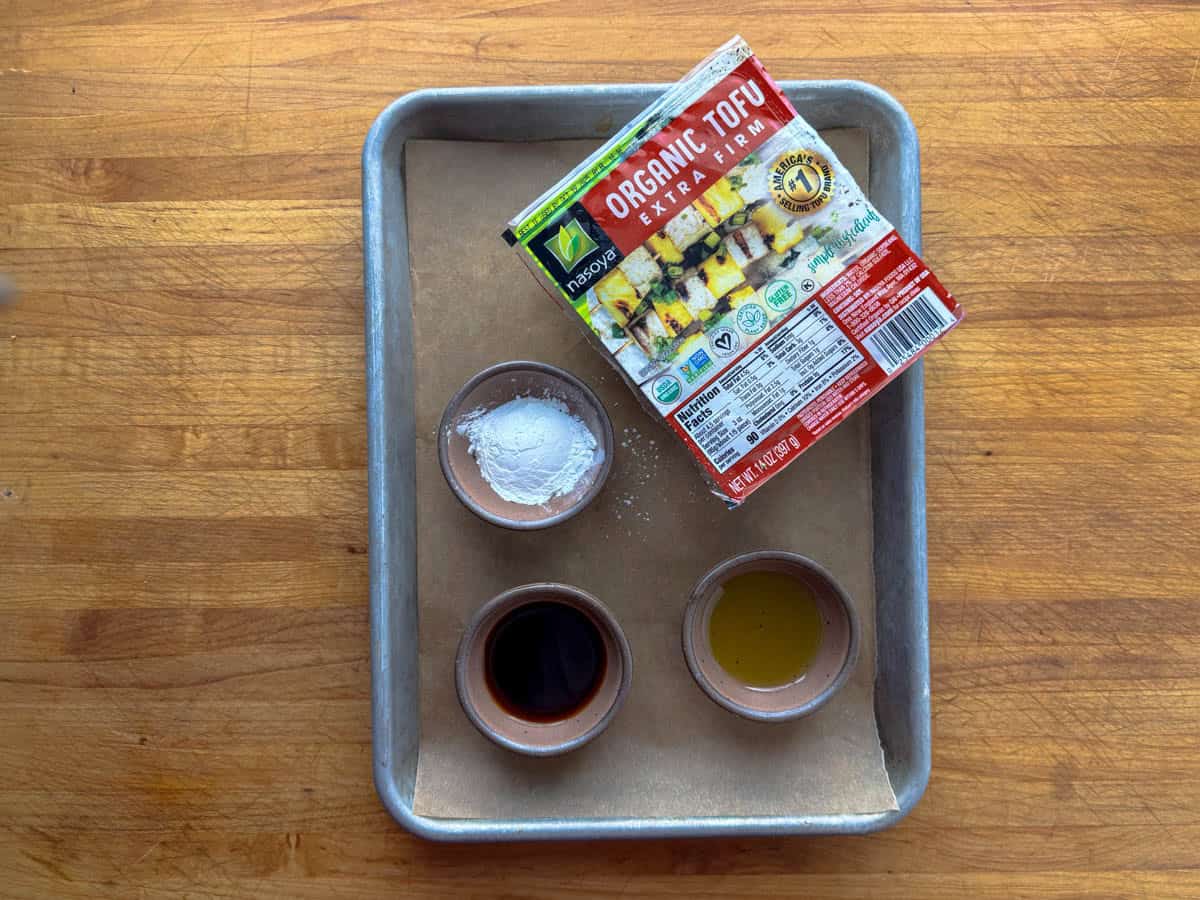 A tray lined with parchment paper holds four small bowls of cornstarch, soy sauce, oil, and maple syrup next to a package of organic extra firm tofu on a wooden surface.