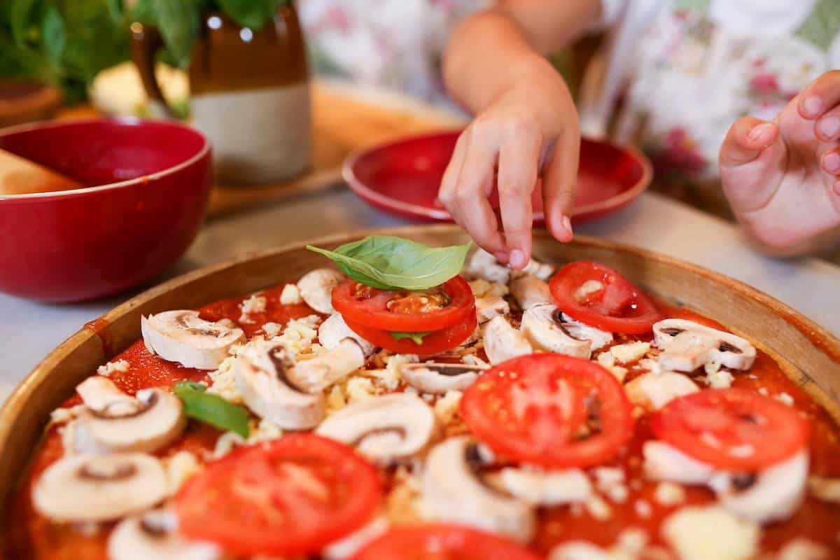 A child's hands add vegetables toppings to a homemade pizza with sliced tomatoes, mushrooms, cheese, and basil, surrounded by red bowls, stovetop cooked apples, and other kitchen items on the table.