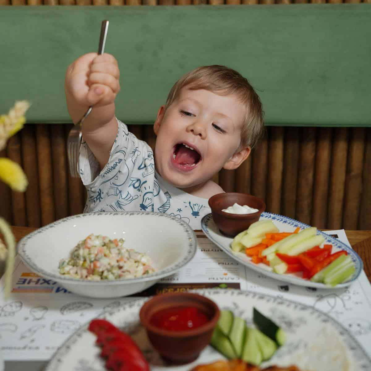A young boy excitedly raises a spoon above a table set with plates of salad, vegetable sticks, stovetop cooked apples, and sauces, smiling with his mouth open in anticipation.
