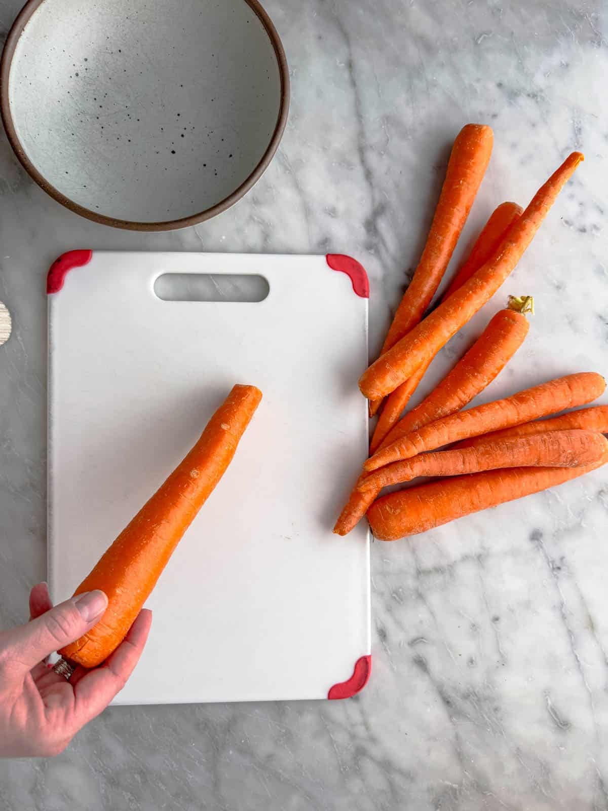 A hand holds a carrot over a white cutting board with several whole carrots on the side, next to a speckled bowl, all on a marble countertop.