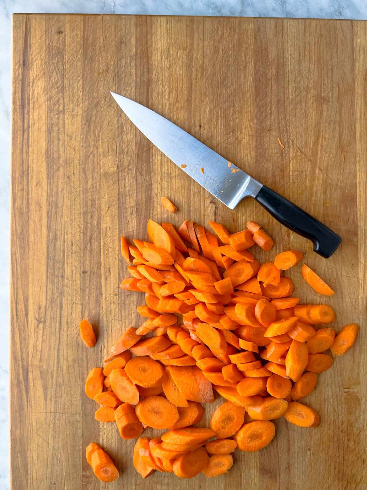 A pile of sliced carrots and a chef’s knife with a black handle are on a wooden cutting board.