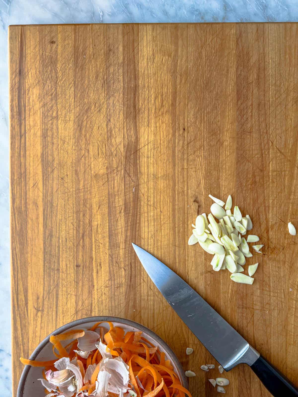 A wooden cutting board with a knife, a small pile of sliced garlic, and a bowl containing garlic skins and carrot peels on a marble countertop.