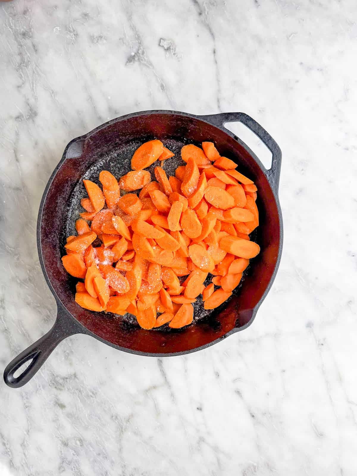 A cast iron skillet filled with sliced raw carrots and a sprinkle of salt sits on a light marble countertop.