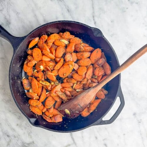 A cast iron skillet filled with cooked, sliced carrots seasoned with herbs, with a wooden spoon resting inside, sits on a light marble countertop.