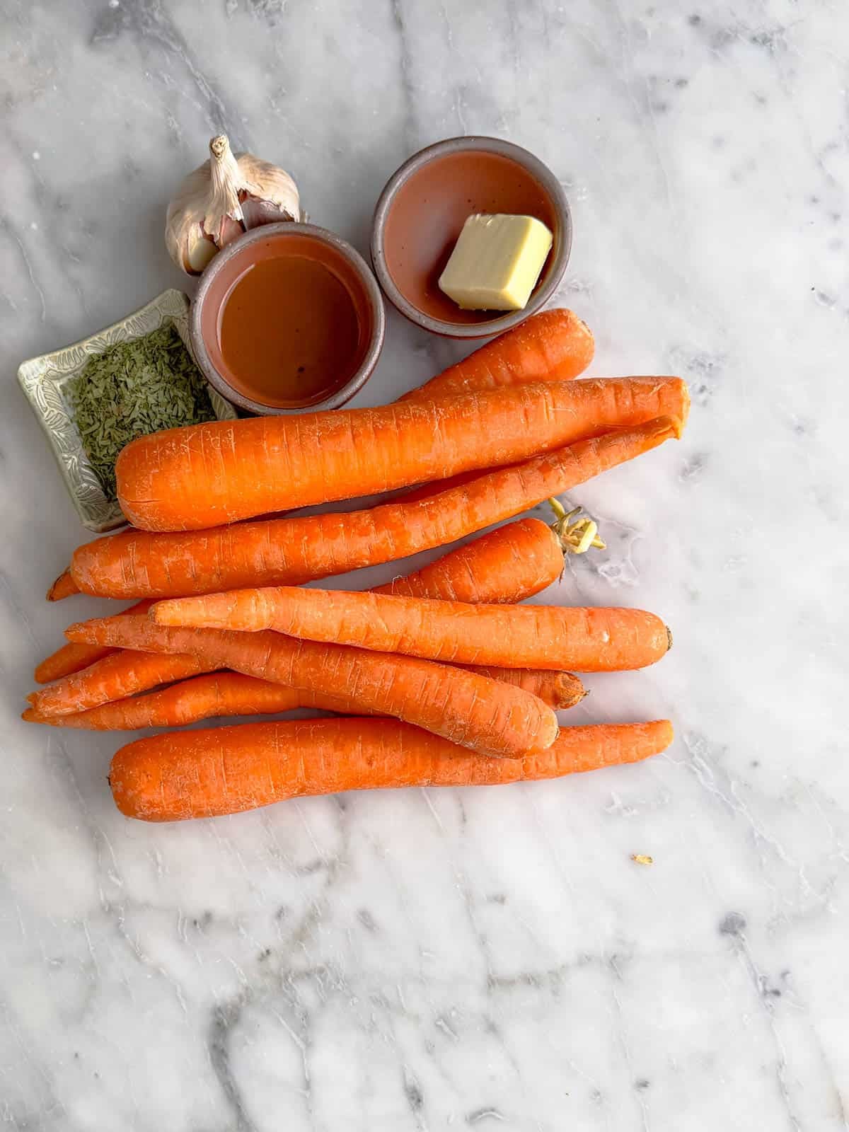 Whole carrots, a square dish of dried herbs, a bulb of garlic, a small bowl of honey, and a small bowl with a piece of butter sit on a light marble surface.