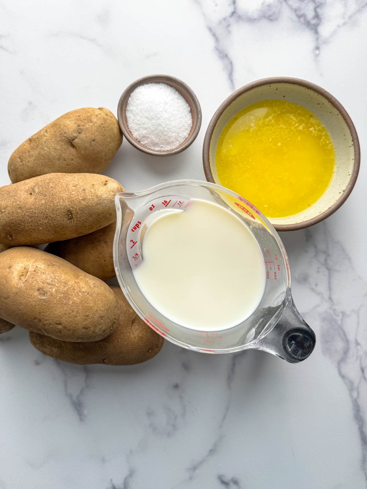Ingredients for easy mashed potatoes: four russet potatoes, a small bowl of salt, a bowl of melted butter, and a measuring cup of milk are arranged on a white marble surface.