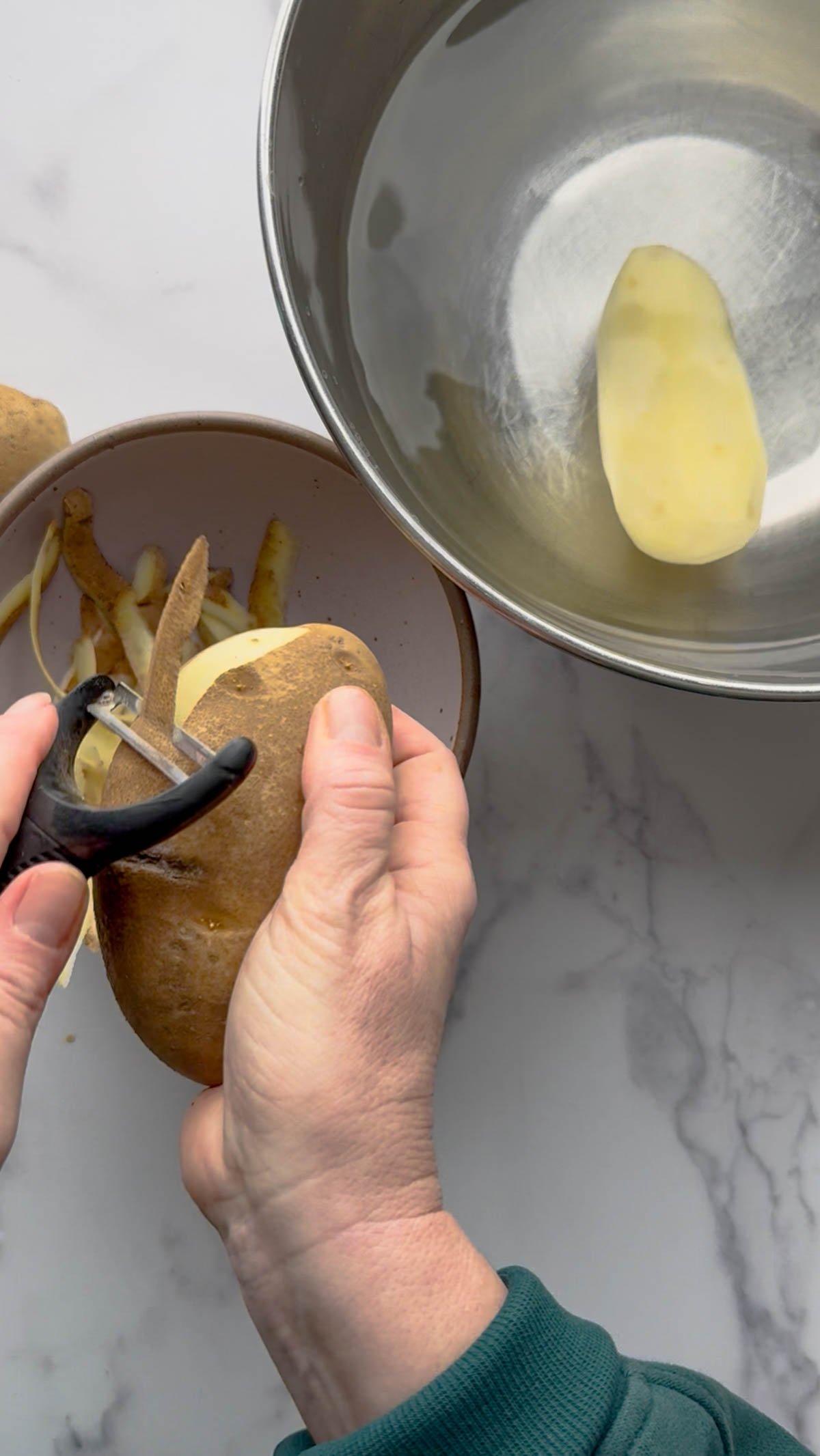 A person peels a potato for mashed potatoes over a bowl, with potato skins collected in one bowl and a peeled slice placed in a metal bowl on a white marble surface.