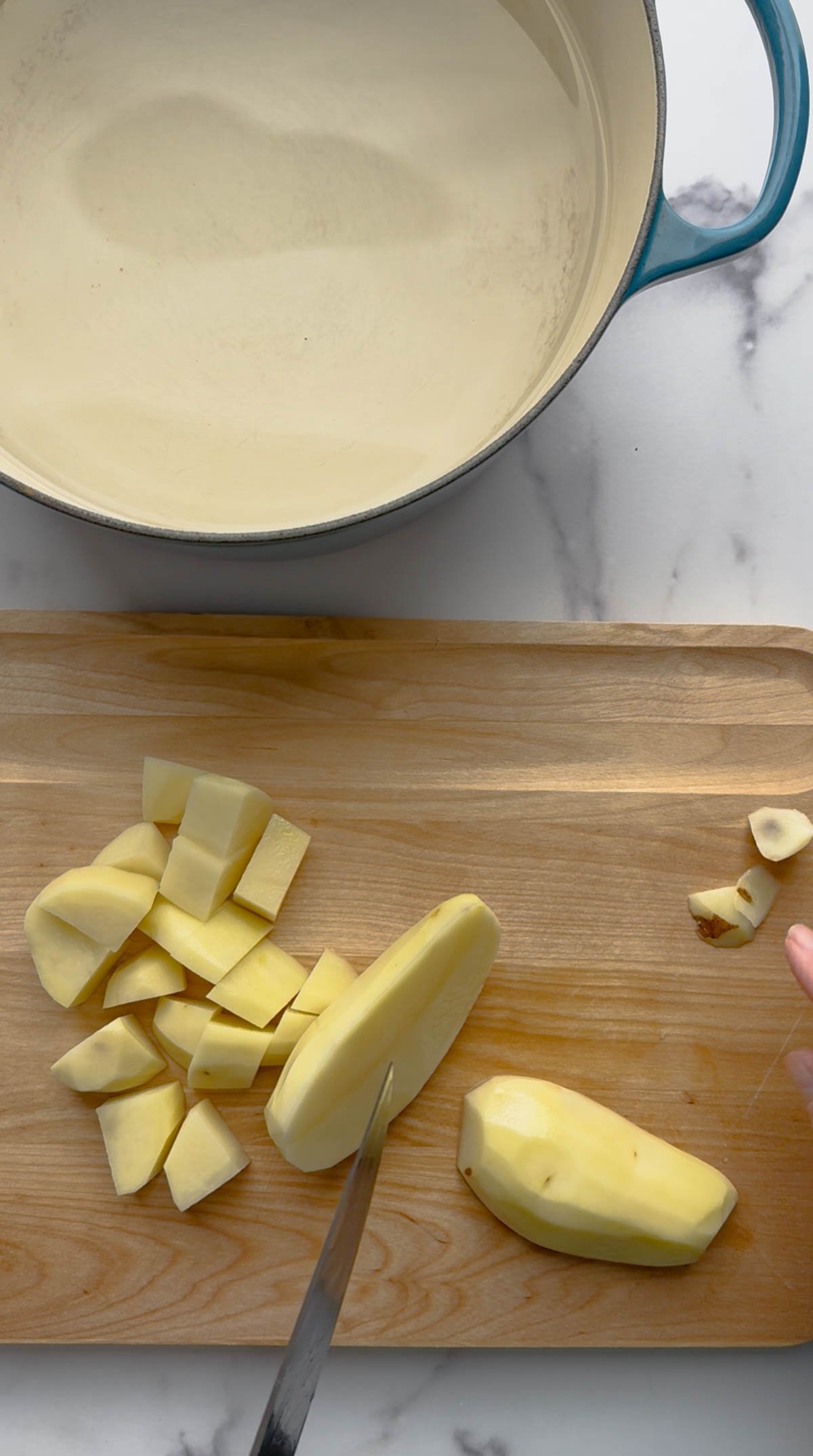 A hand chops peeled potatoes into chunks on a wooden cutting board, with a large cream-colored pot nearby on a marble countertop.