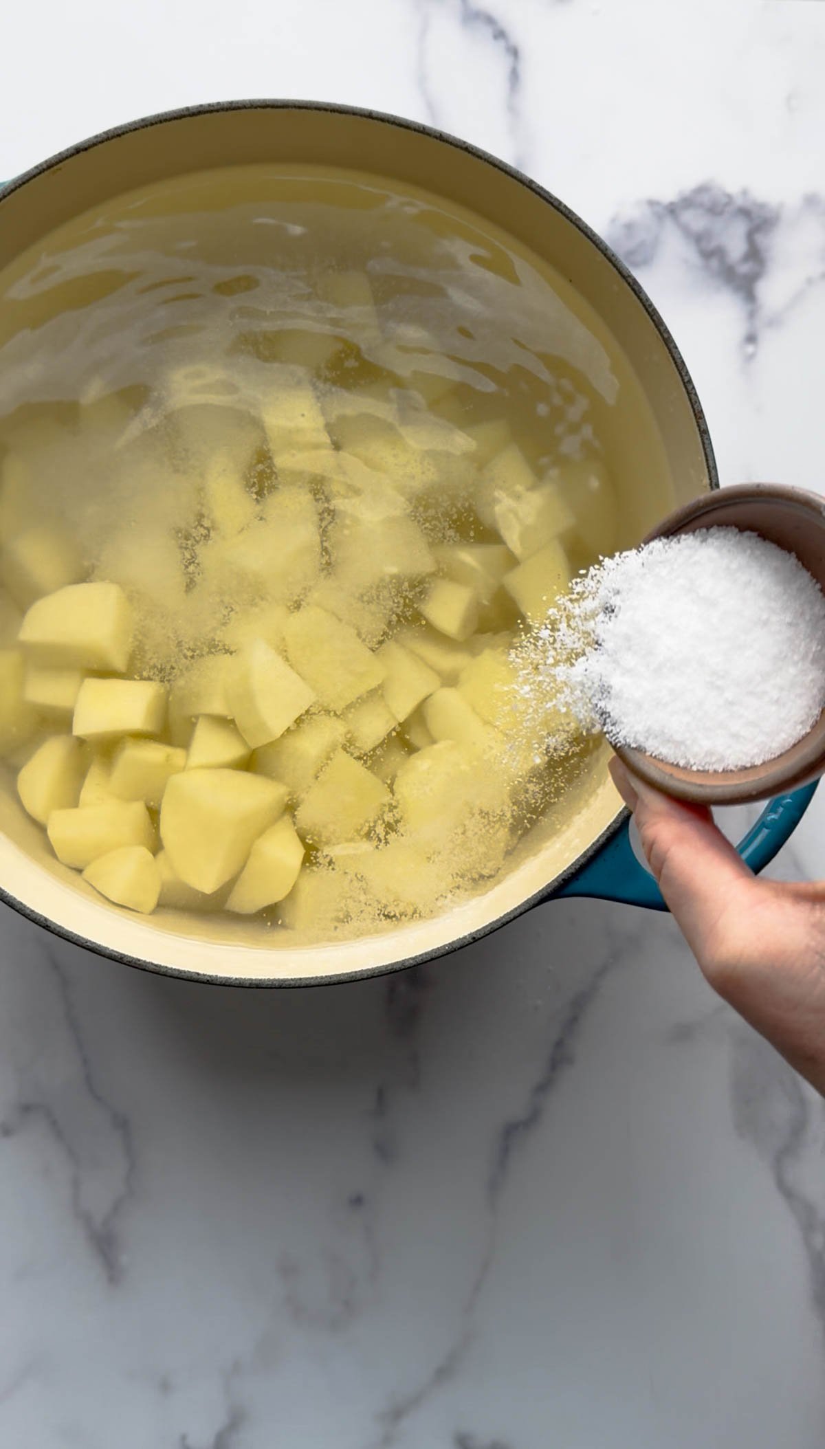 A hand is pouring a bowl of salt into a pot filled with water and peeled, cubed potatoes on a marble countertop.