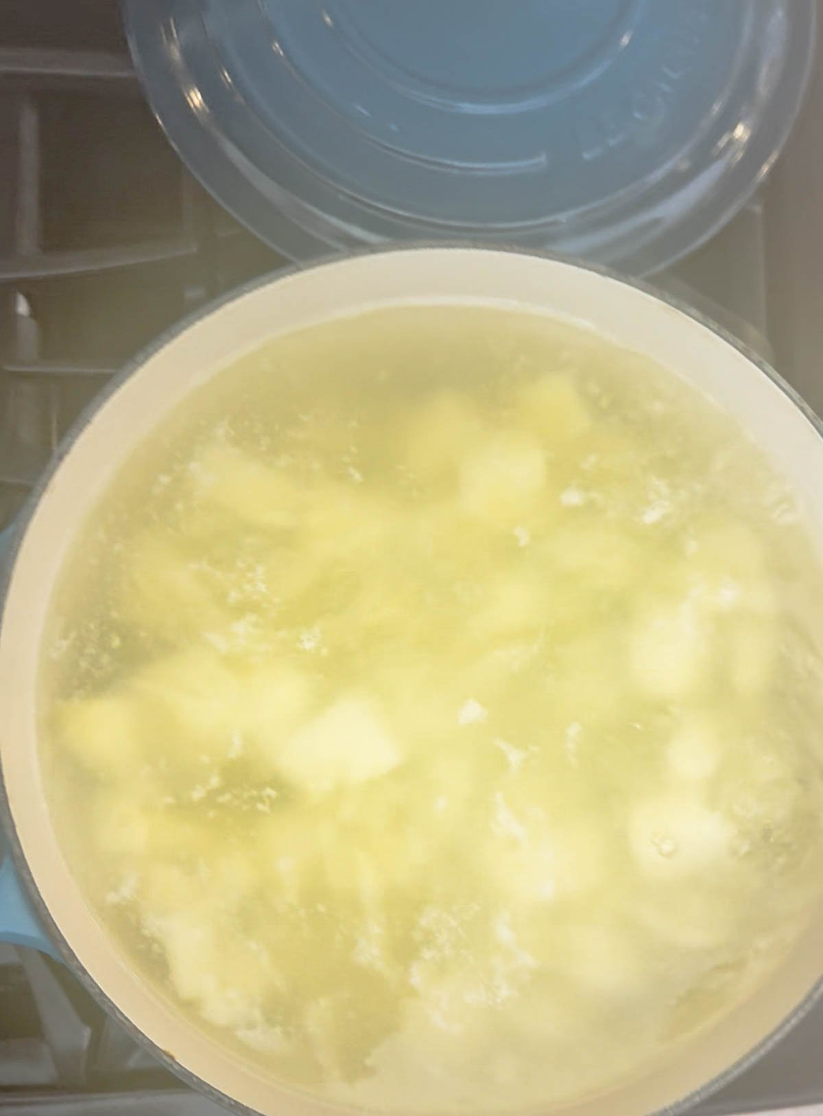 Potato chunks boiling in a light-colored pot filled with water on a stove, with a blue lid placed nearby. Steam is visible, giving the image a slightly hazy appearance.