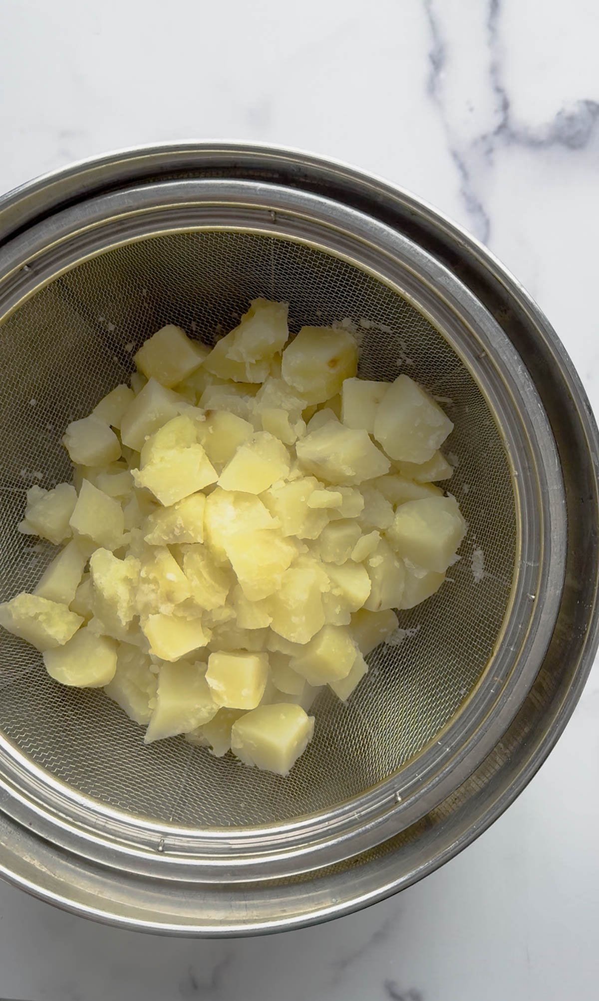 Cubed, cooked potatoes are draining in a metal mesh strainer set over a metal bowl on a white marble surface.