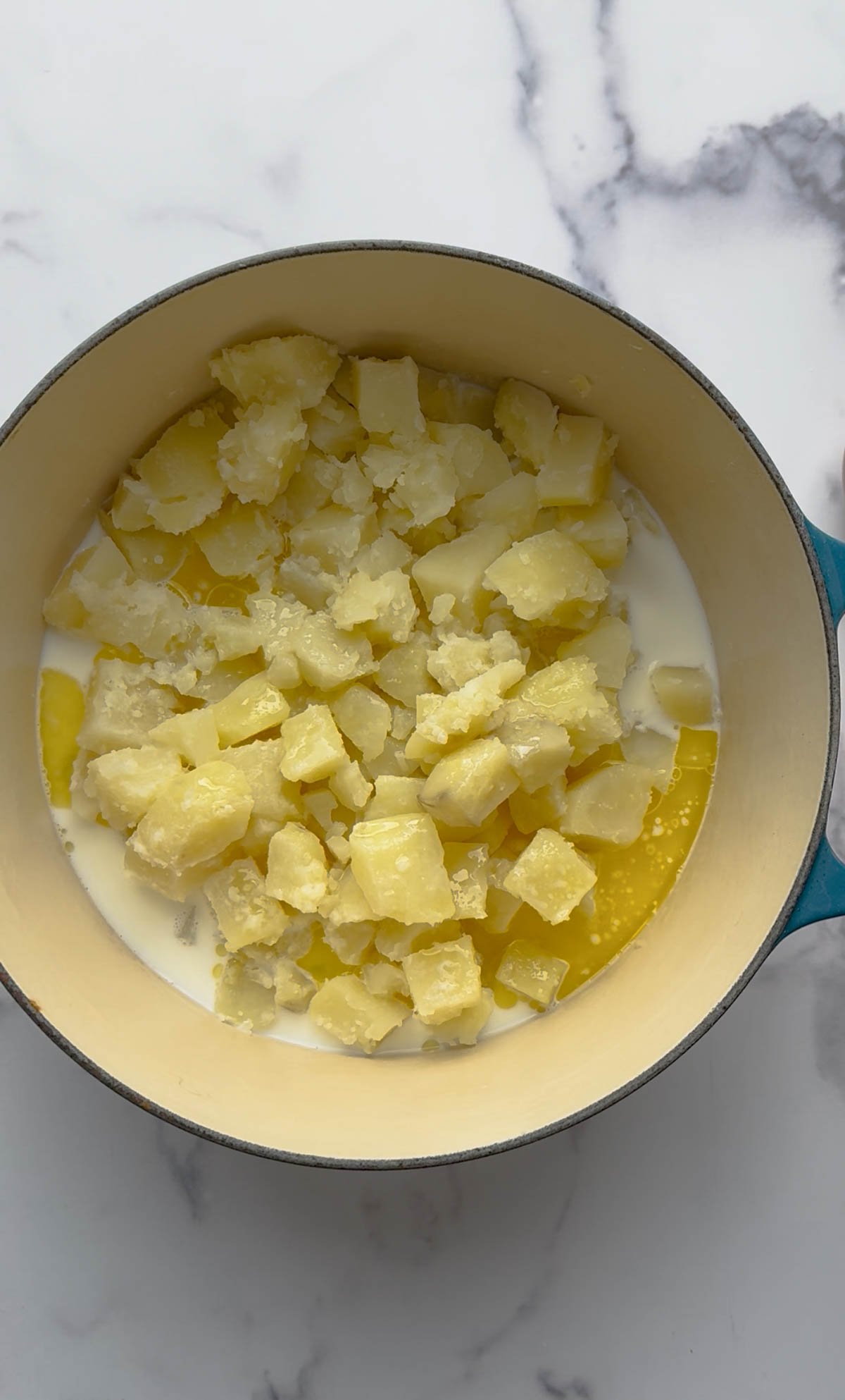 A pot filled with diced cooked potatoes, milk, and melted butter sits on a white marble surface, ready to be mixed.
