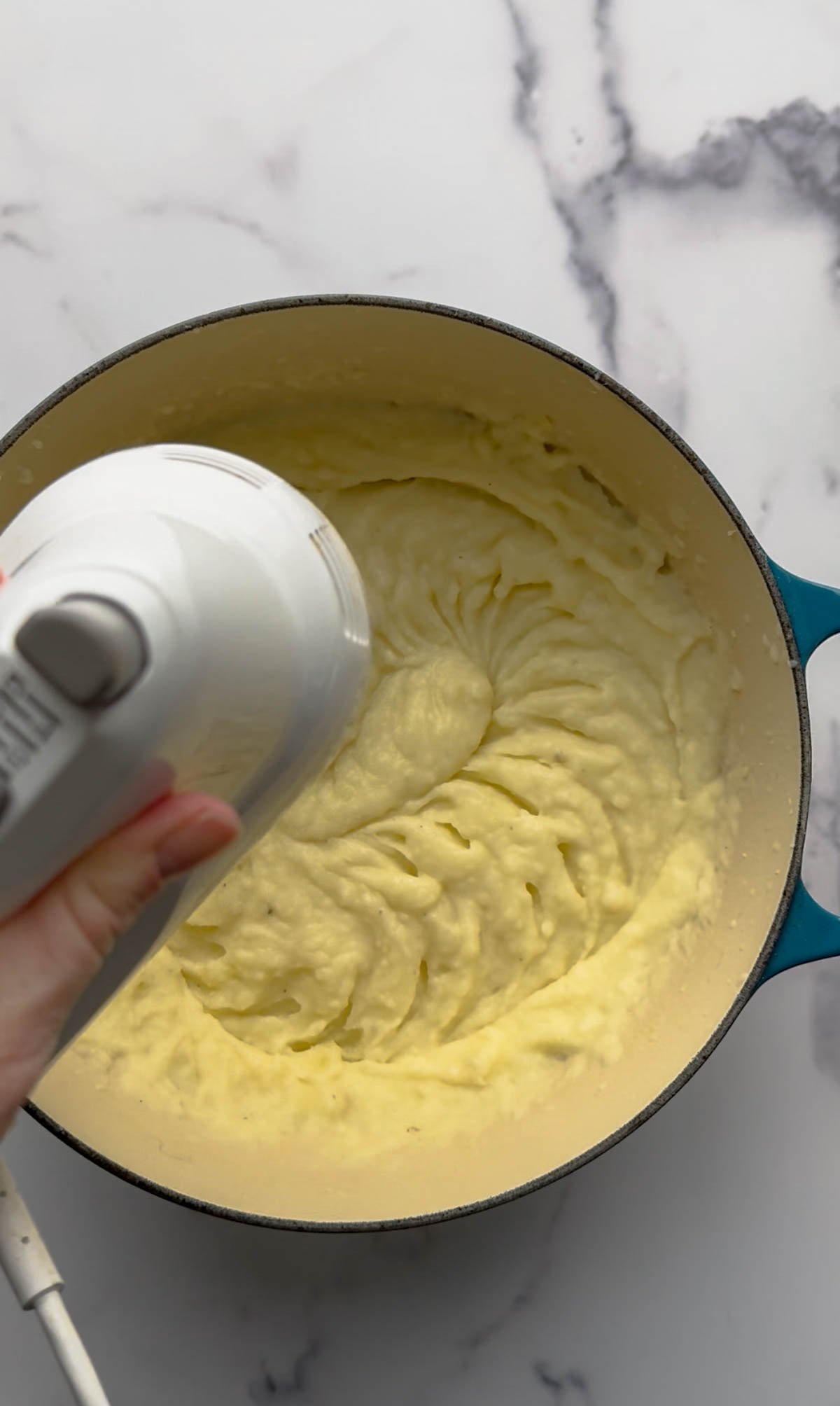 A hand holds a white electric mixer blending creamy mashed potatoes in a blue pot on a white marble countertop.
