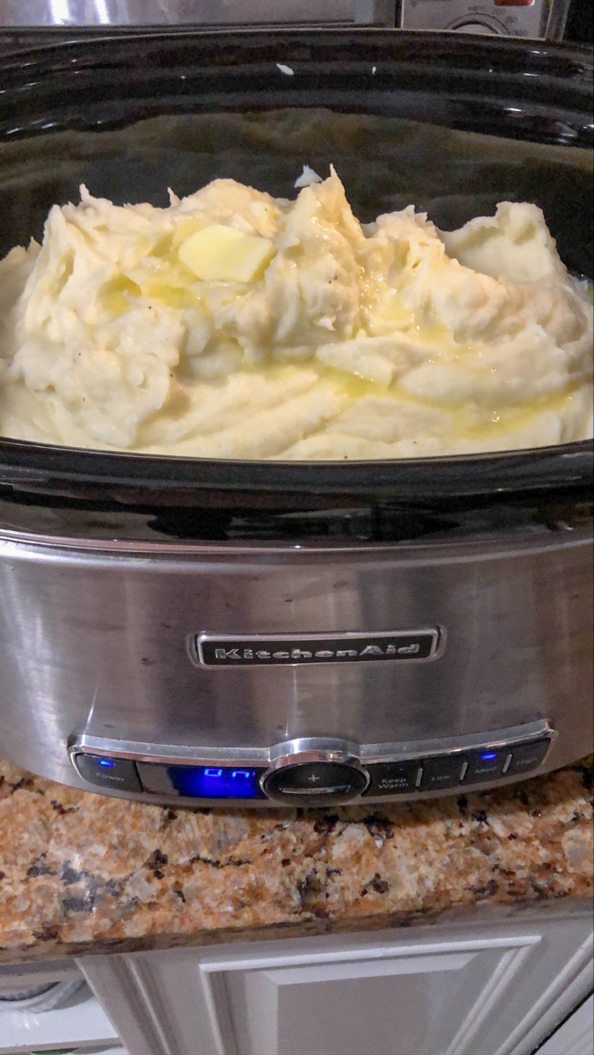 A slow cooker filled with creamy mashed potatoes topped with melted butter sits on a granite countertop. The slow cooker is stainless steel and has the KitchenAid logo on the front.
