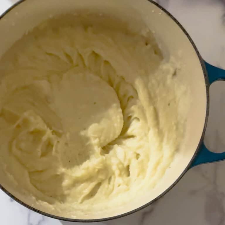 A blue pot filled with creamy, smooth mashed potatoes sits on a white marble surface. The fluffy texture and elegant swirls make this an inviting mashed potatoes recipe display.