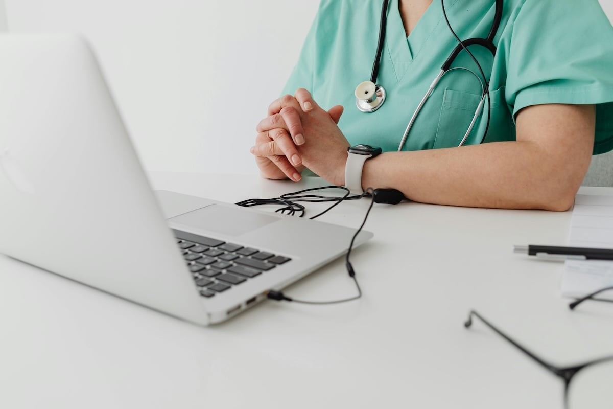 A healthcare professional in green scrubs with a stethoscope sits at a desk, hands clasped, facing an open laptop. A medical document about interpreting growth charts and a pair of eyeglasses are neatly placed on the desk.