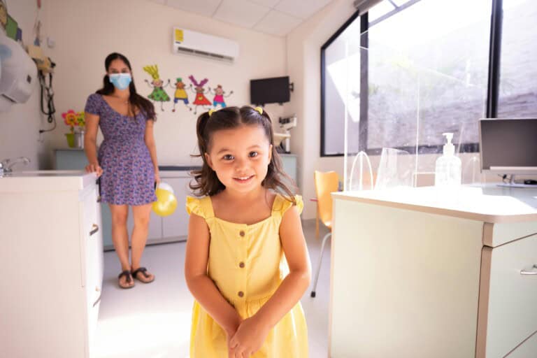 A young girl in a yellow dress smiles at the camera in a brightly lit doctor's office, while a woman in a mask and patterned dress stands by the counter, possibly interpreting growth charts in the background.