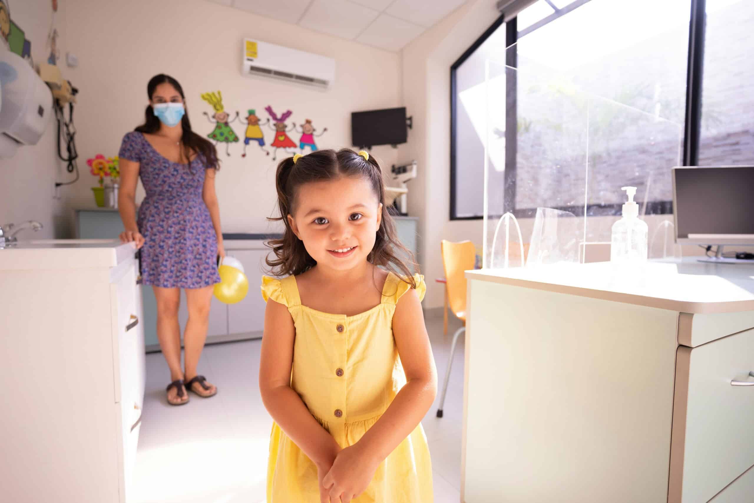 A young girl in a yellow dress smiles at the camera in a brightly lit doctor's office, while a woman in a mask and patterned dress stands by the counter, possibly interpreting growth charts in the background.