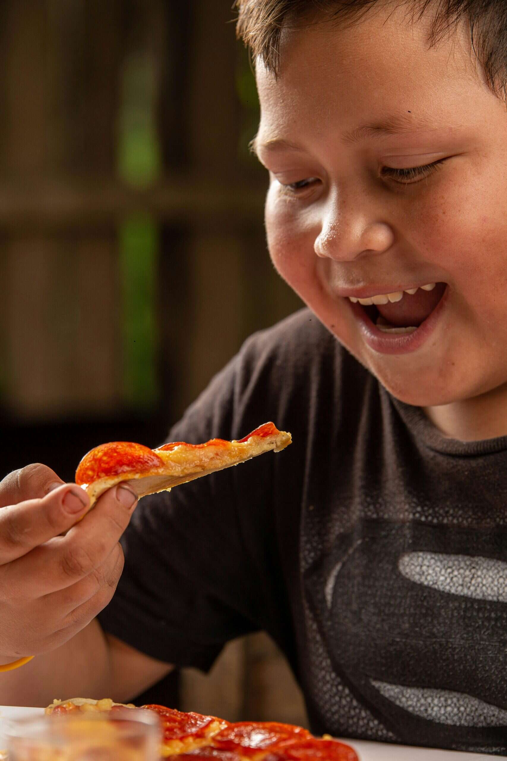 A young boy smiles excitedly while holding a slice of pepperoni pizza, ready to take a bite. Wearing a dark shirt and sitting at a table with more pizza, he looks as cheerful as when he’s interpreting growth charts in class.