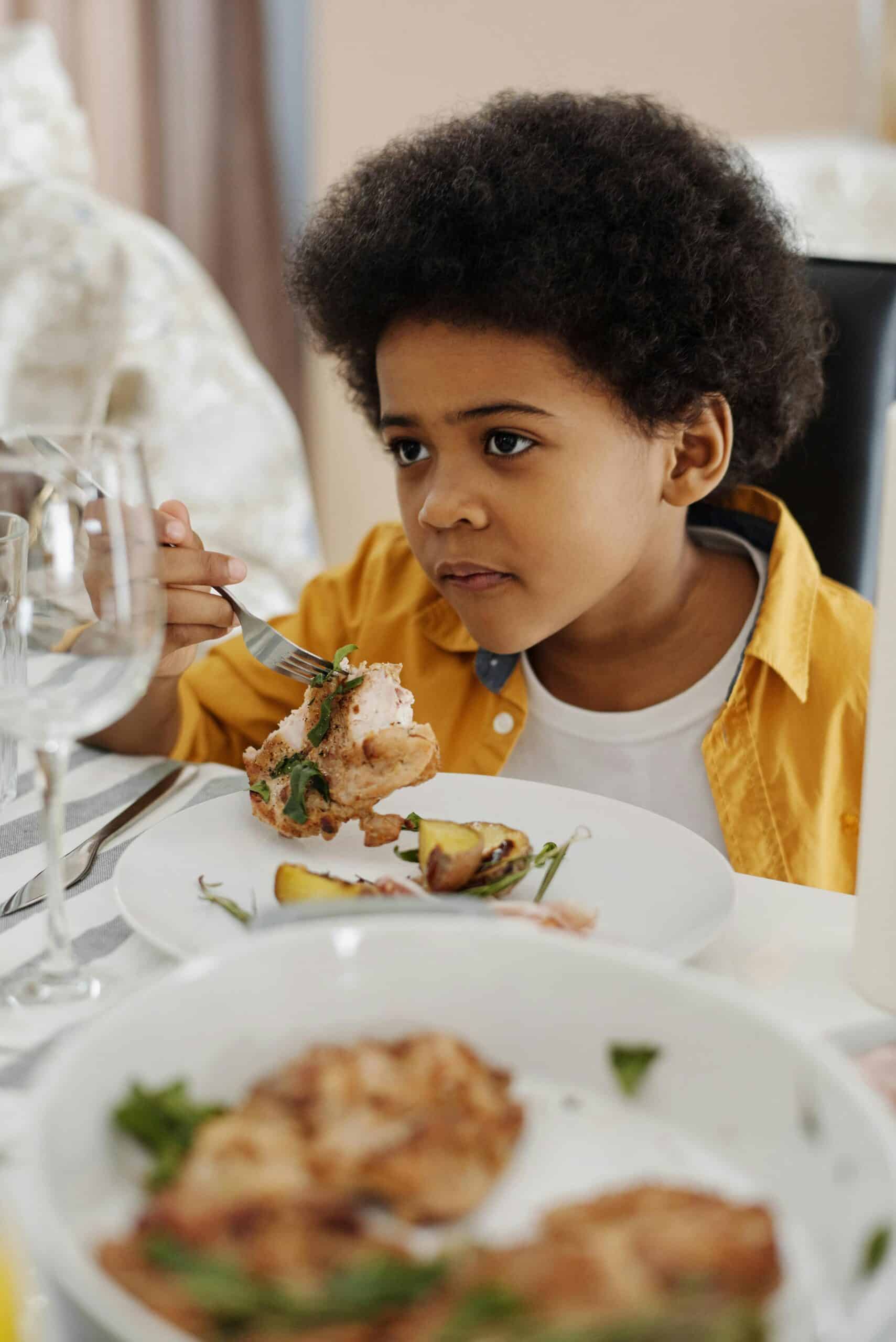 A young child with curly hair, wearing a yellow shirt, sits at a dining table holding a fork with food, looking thoughtful—perhaps as if interpreting growth charts while surrounded by plates of food and glasses.