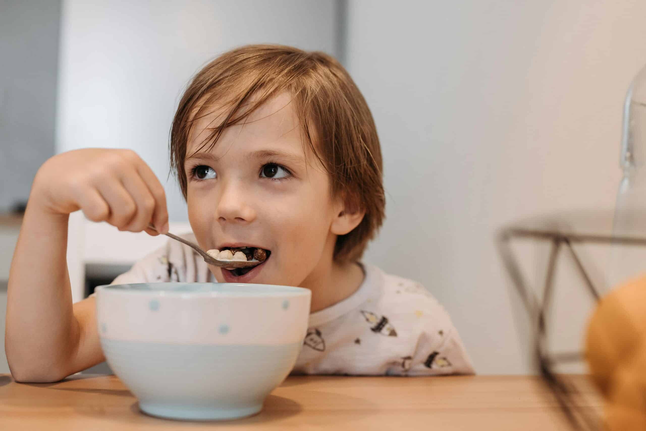 A young child with light brown hair eats cereal from a white bowl with a spoon, smiling and looking up while sitting at a wooden table, as a parent nearby considers interpreting growth charts for healthy development.